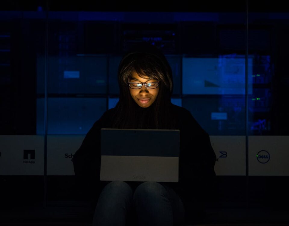 Link dedicado e logística: otimização nas operações online A woman uses her laptop in a dimly lit server room, focusing on technology and work.