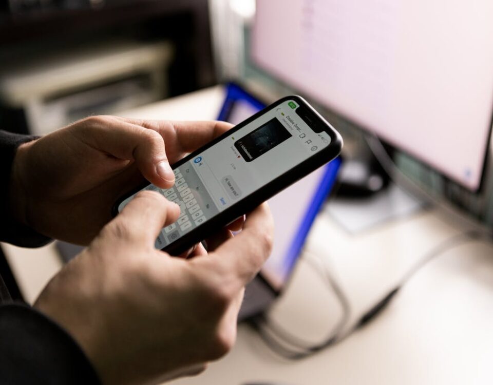 Como robôs ajudam na gestão de pedidos via Whatsapp A person holding a smartphone and chatting indoors near a computer screen.