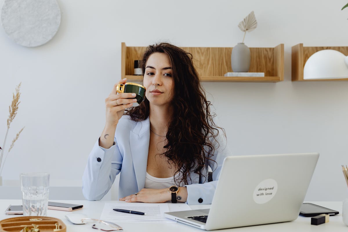 Gestão de estoque que melhora o e-commerce Confident businesswoman enjoying a cup of coffee while working on her laptop in a chic office setting.