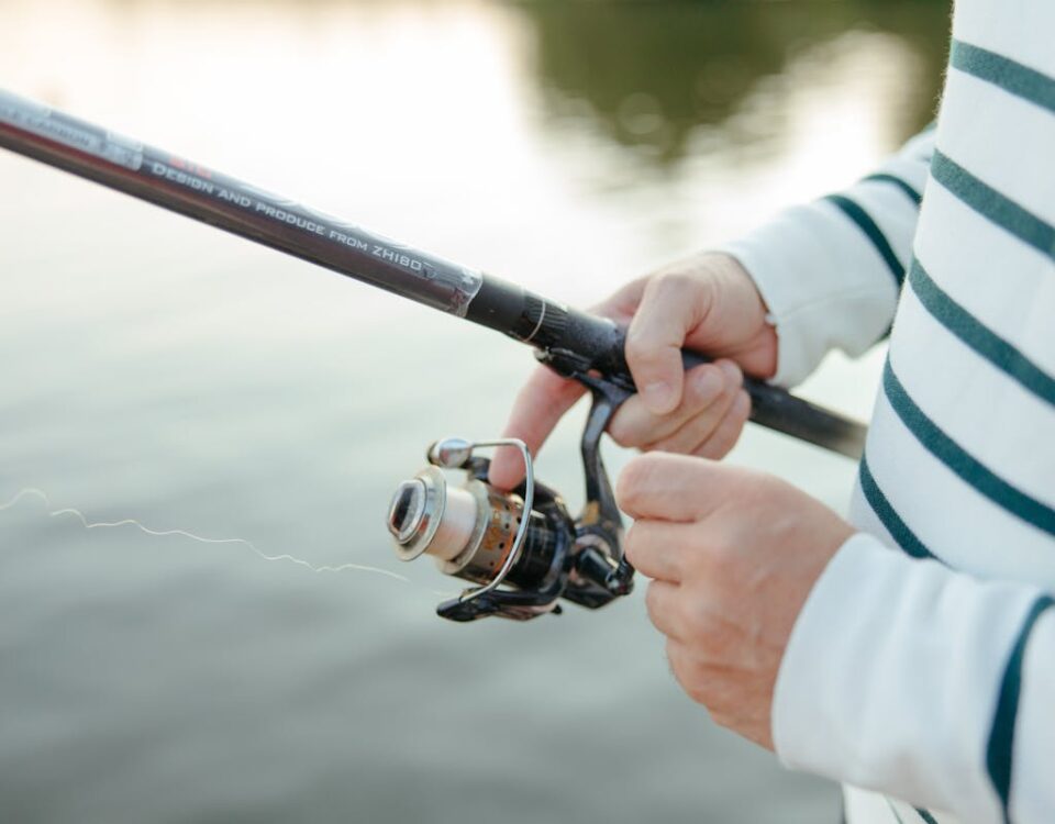 Onde comprar equipamentos de pesca online com segurança e confiança Close-up of hands holding a fishing rod with a wheel by the water, capturing leisure and recreation.