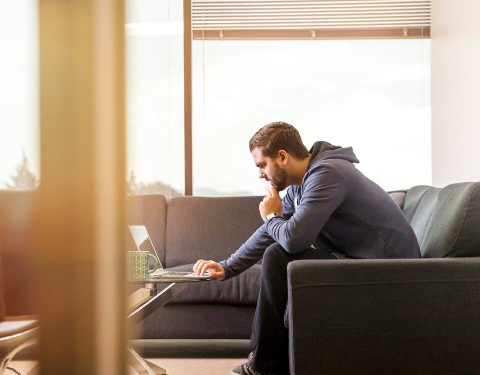 Logística para auxiliares administrativos: um guia prático A man in casual attire works remotely from a sofa, using a laptop near a large window in a modern room.