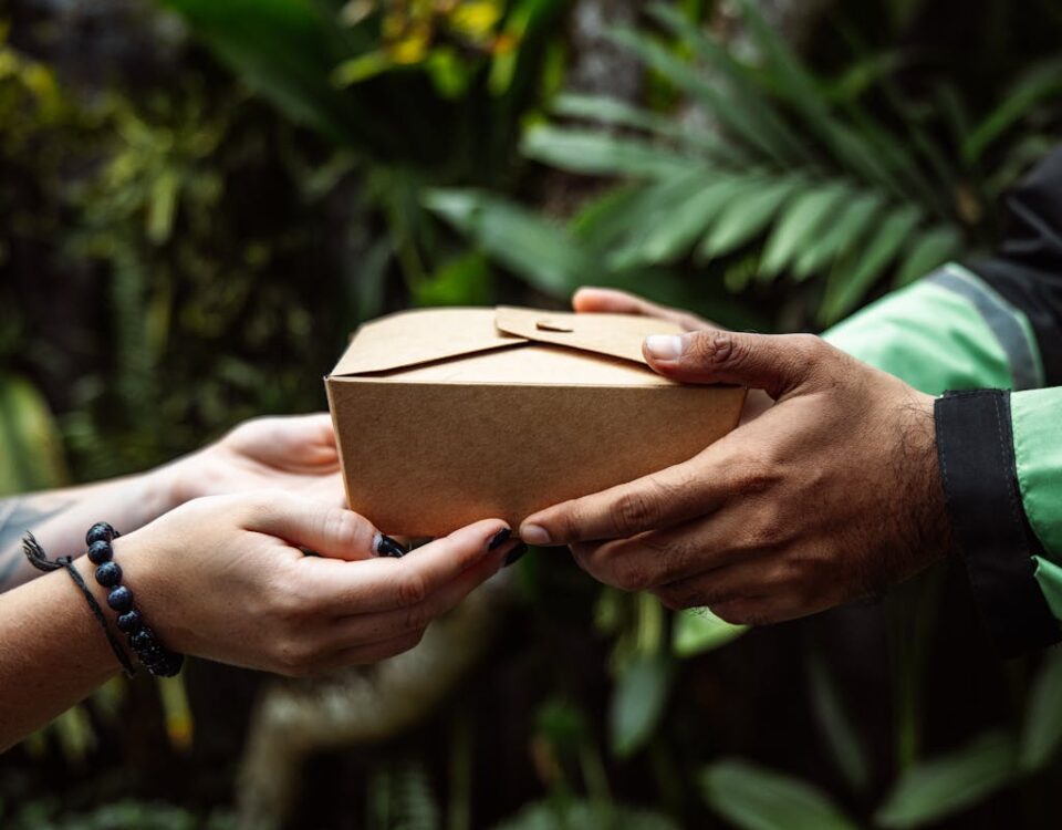 Estratégias logísticas para o mercado adulto Close-up of hands exchanging a cardboard package in a lush green setting, representing eco-friendly delivery.