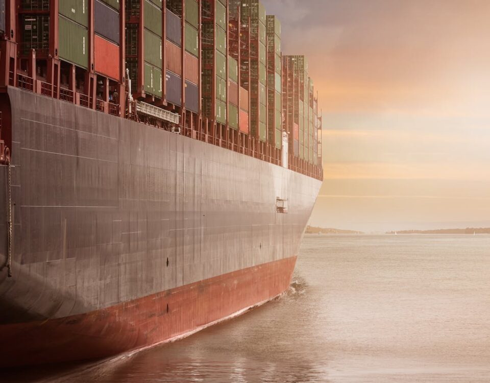 A colossal cargo ship loaded with containers navigates through calm waters against a vibrant sunset sky.
