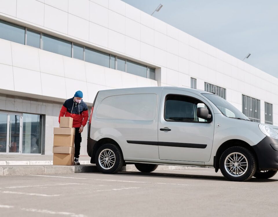 Seguros para veículos de entrega: Vale a pena investir? A delivery driver wearing a face mask unloads packages from a van outside a modern building.