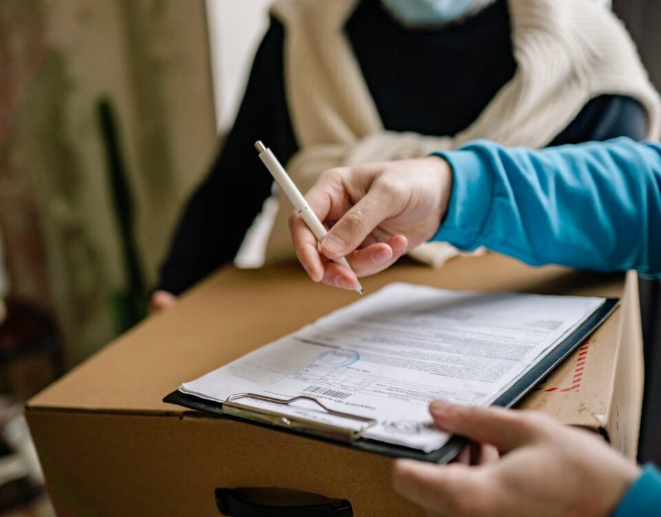 É possível comprar carabinas online com segurança? Close-up of a person signing a delivery document placed on a large cardboard box indoors.