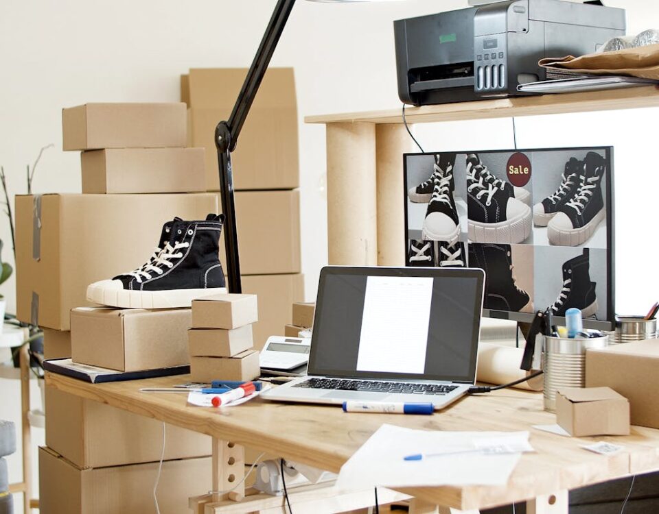 Painéis SMM ajudam no e-commerce? A busy workspace with sneakers, boxes, and a laptop indicating an online shoe store or e-commerce setup.