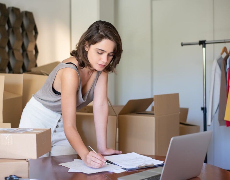 Automação comercial: Impacto direto no e-commerce Young woman managing her online clothing business from home office with boxes and laptop.