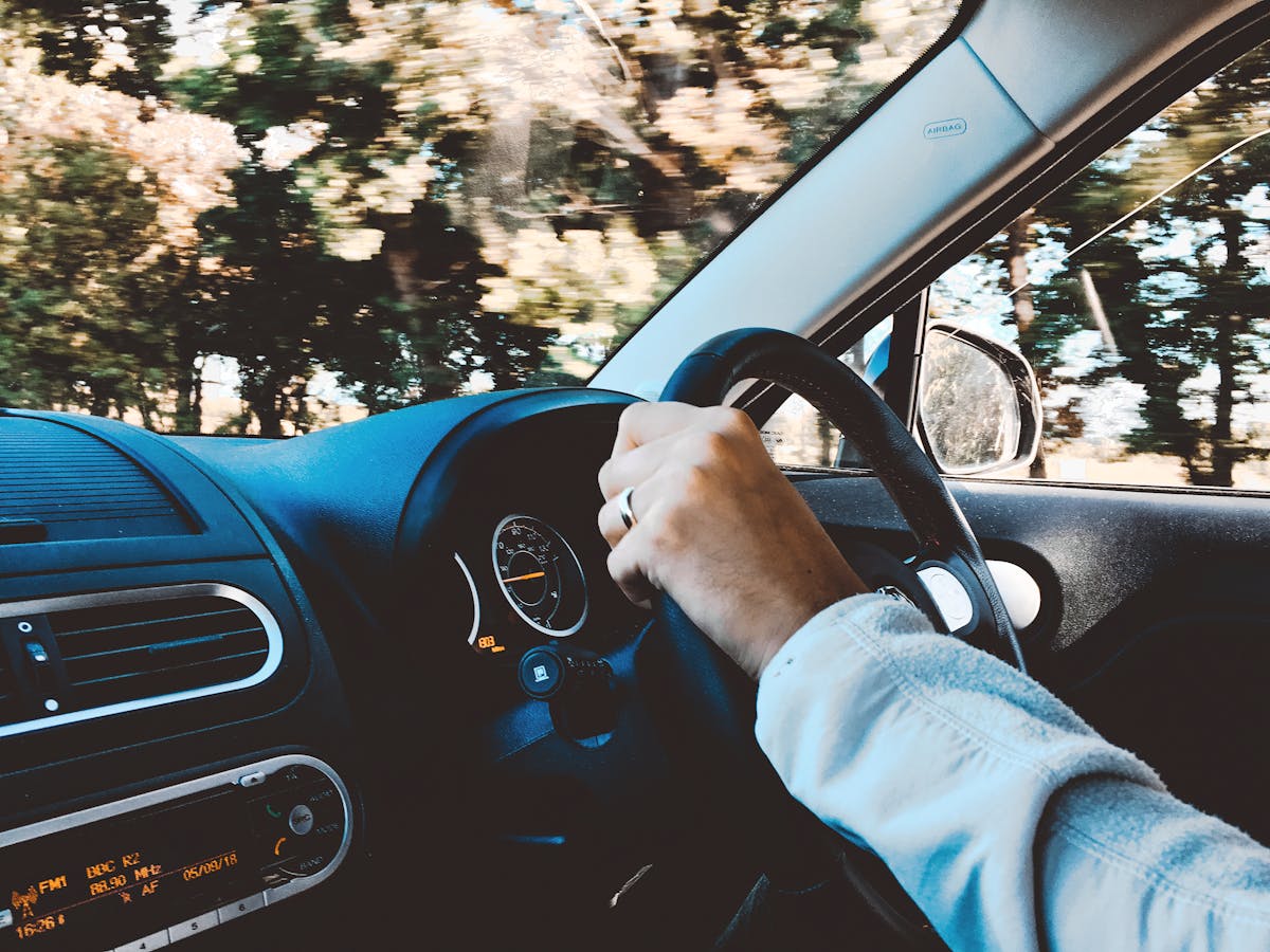 Direção econômica: um diferencial no mercado de entregas Driver's hand on steering wheel of a modern car during a daytime drive through scenic outdoors.