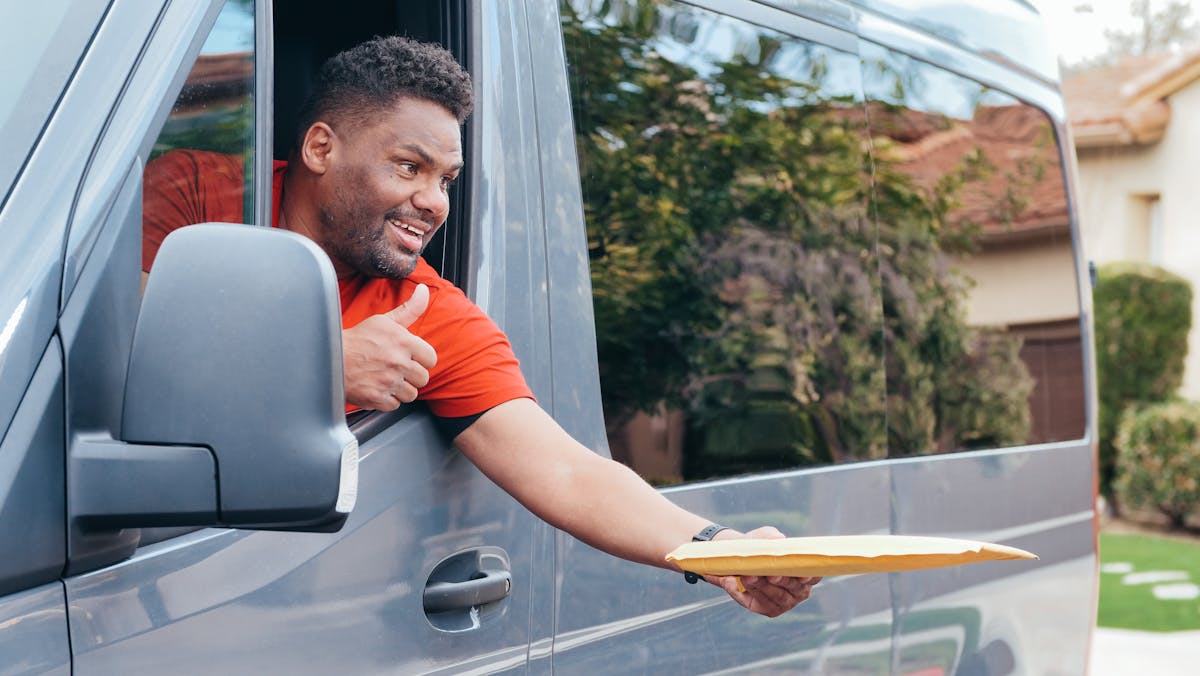 A delivery driver in a red shirt gives a thumbs up while handing a parcel from a van.