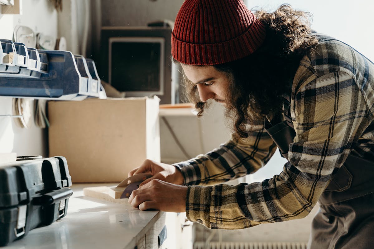 Focused young male carpenter sanding wood at a workbench indoors.