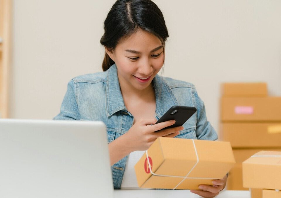 Lojista conferindo pedido no celular ao lado de notebook e caixas de papelão